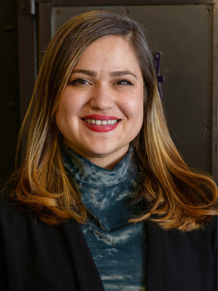 Smiling person with straight shoulder-length brown hair, wearing a dark blazer and a blue patterned scarf