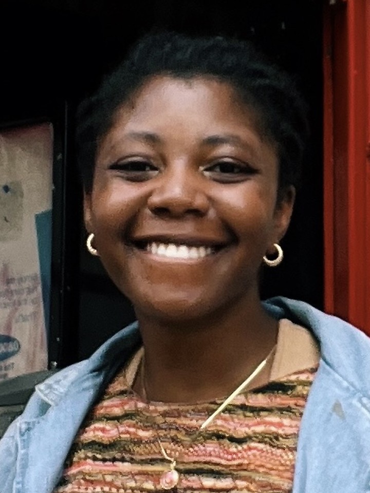 Smiling Black woman with small hoop earrings, a patterned shirt under a collared jean shirt, and a necklace