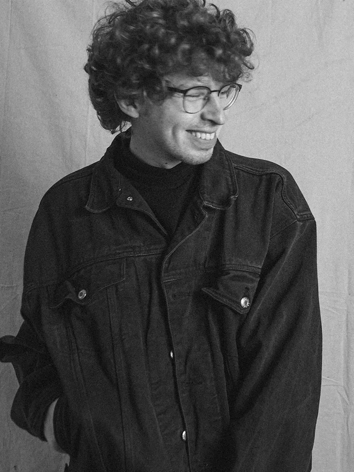 A black and white photo of Ryan Shimko, a young man with curly hair, wearing a dark jean jacket and glasses. Ryan is smiling while looking to the side.