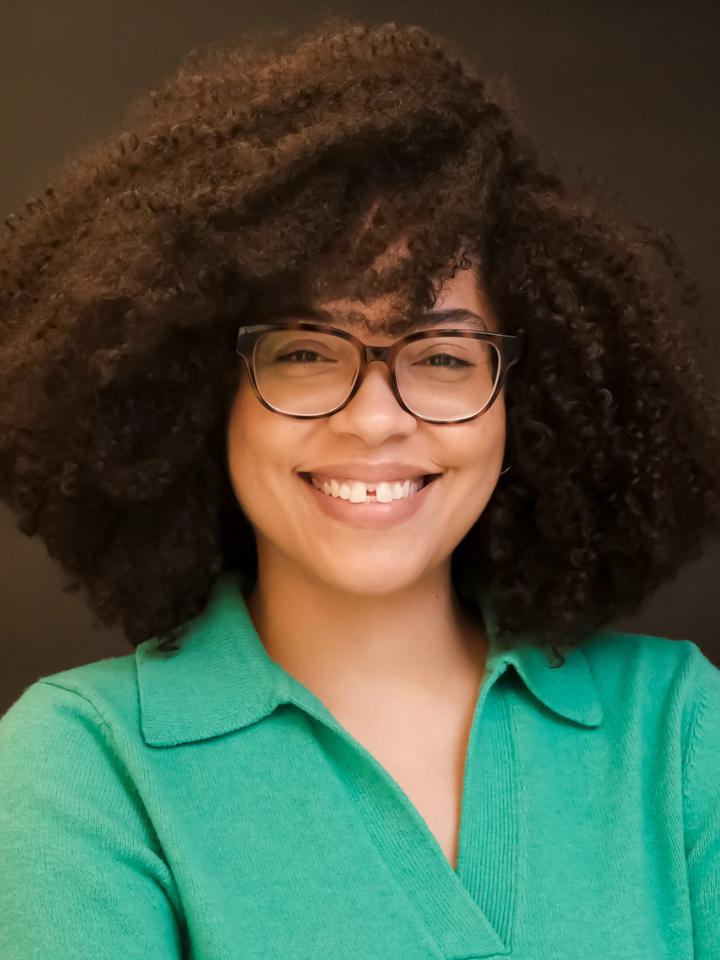 Smiling person with curly dark hair and rectangular glasses, wearing a teal green collared top