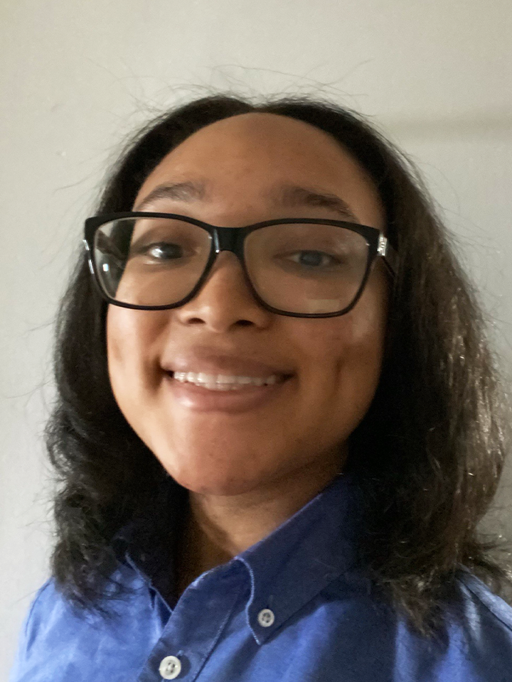An image of Trinity Spencer, a young black woman with black glasses and short black hair. She is smiling, with a blue collared shirt on.