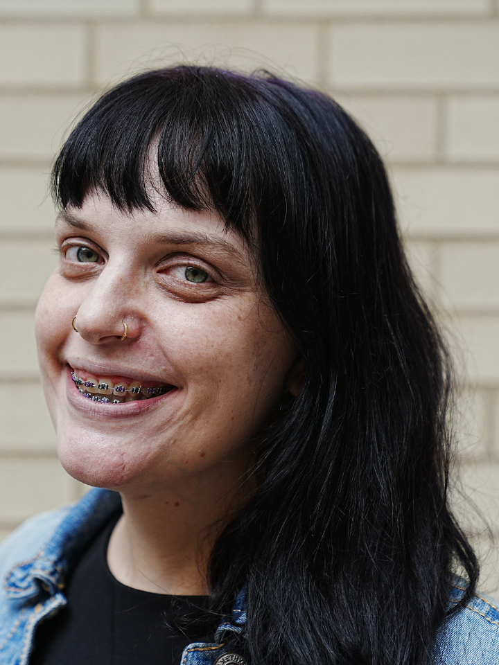 Close-up portrait of a person with long dark hair with nose rings and braces, wearing a jean jacket over a black shirt