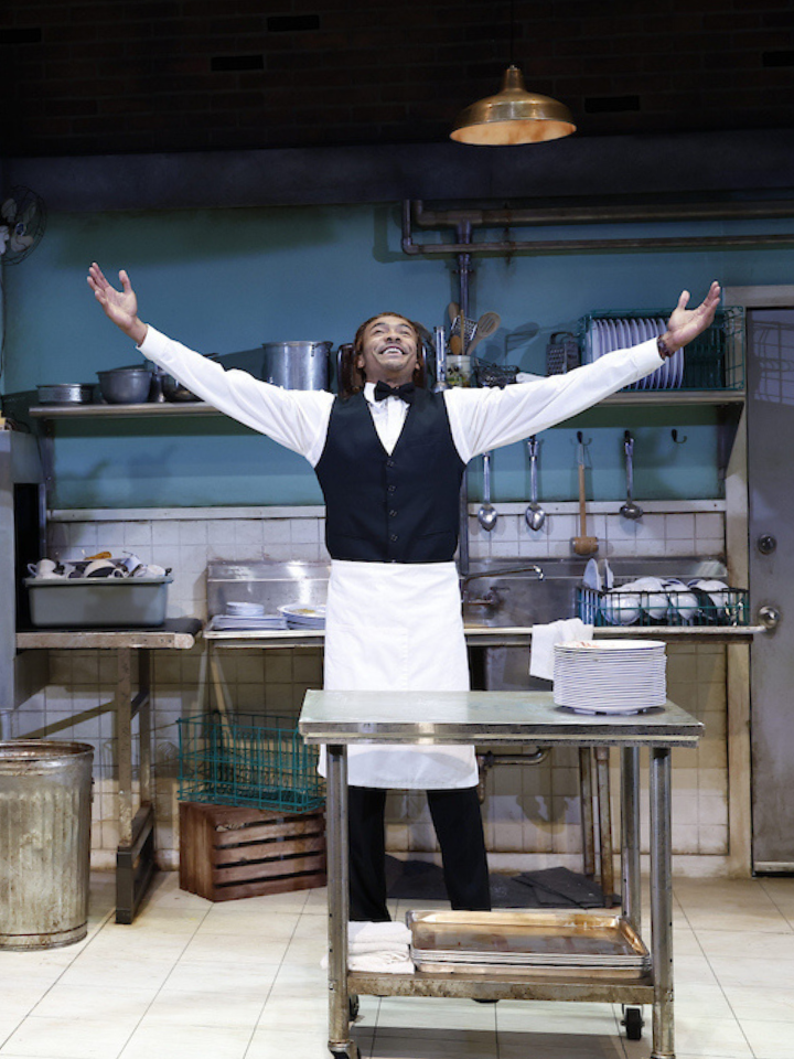 A man stands in a restaurant kitchen with arms raised dramatically.