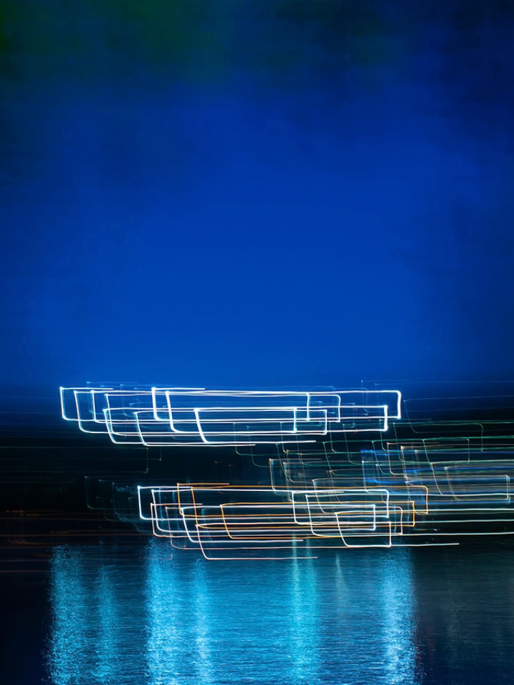 Long exposure of light trails reflected on water against a dark blue background.