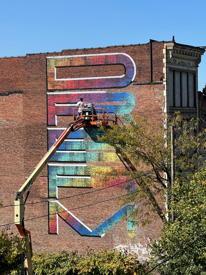 Two people in a lift paint a colorful, large-lettered mural on a brick building wall.