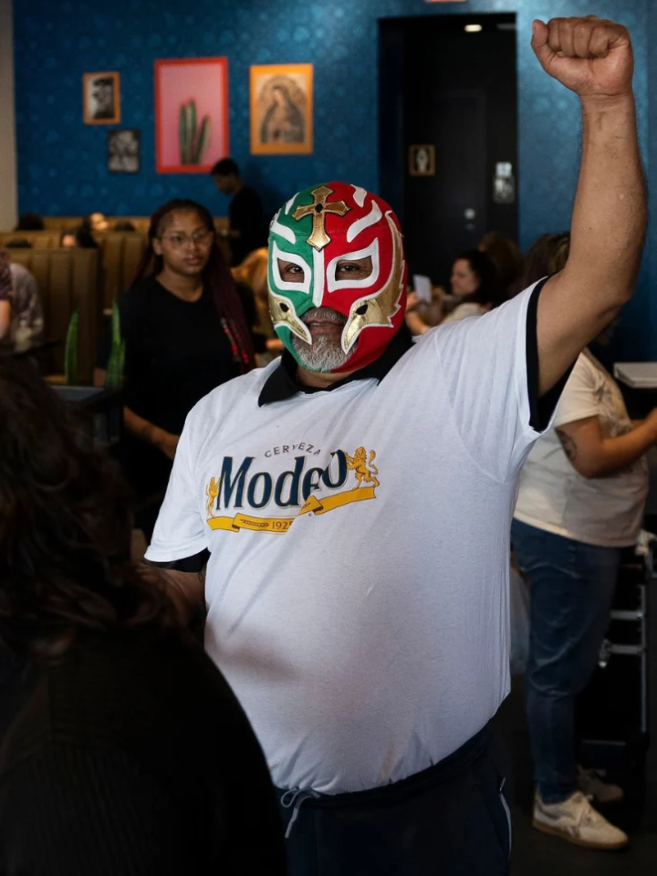 A man wearing a luchador mask and a Rodeo beer t-shirt raises his fist in a crowded restaurant.