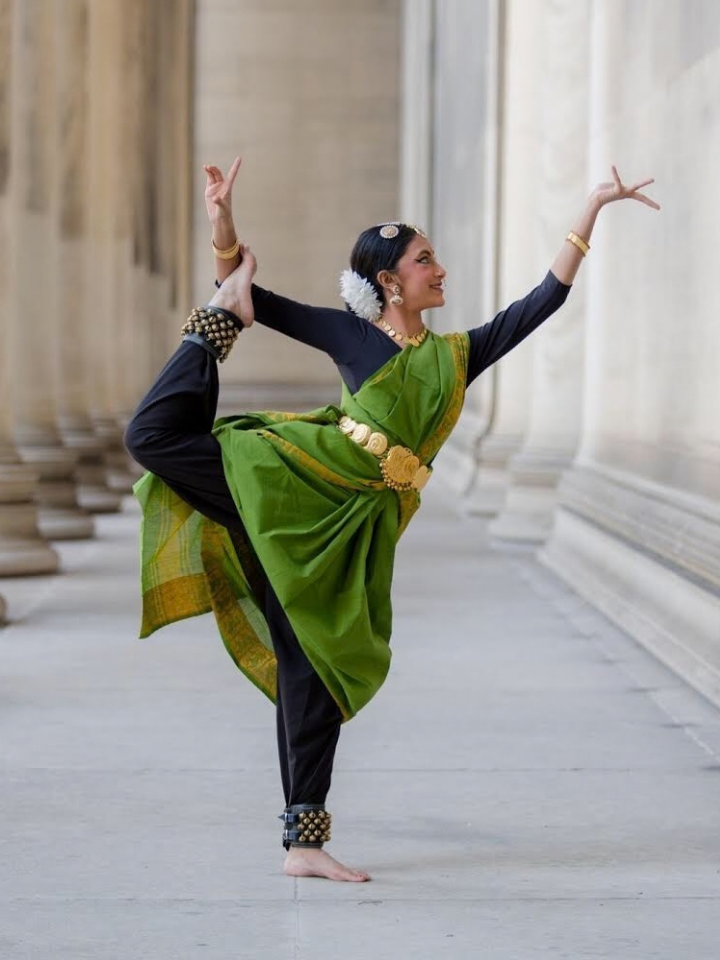 A woman in green Indian traditional attire poses in a classical dance stance between tall columns.