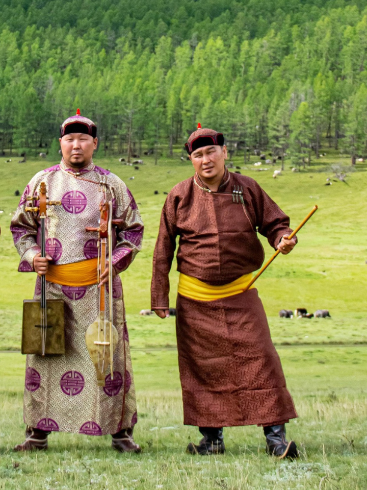 Three men in traditional Mongolian clothing stand on a grassy field with trees and grazing animals in the background. One holds a stringed instrument, and all wear hats and bright yellow sashes.