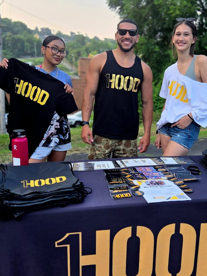 Three people stand behind a table of HOOD branded apparel and materials.