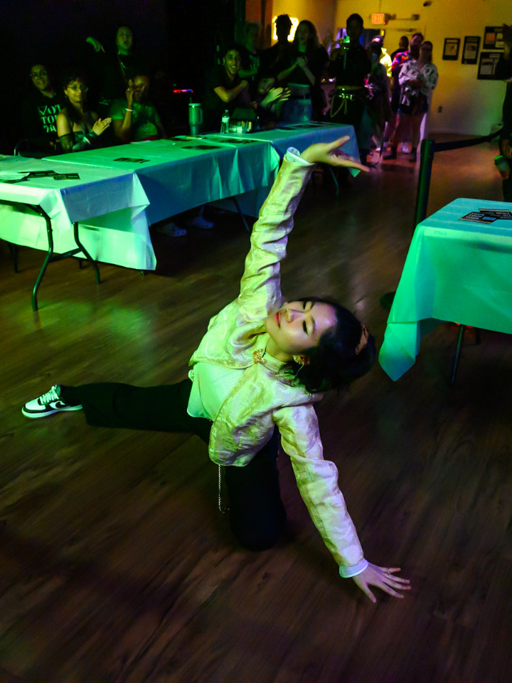 A person poses on a wooden floor in front of tables at an indoor event.