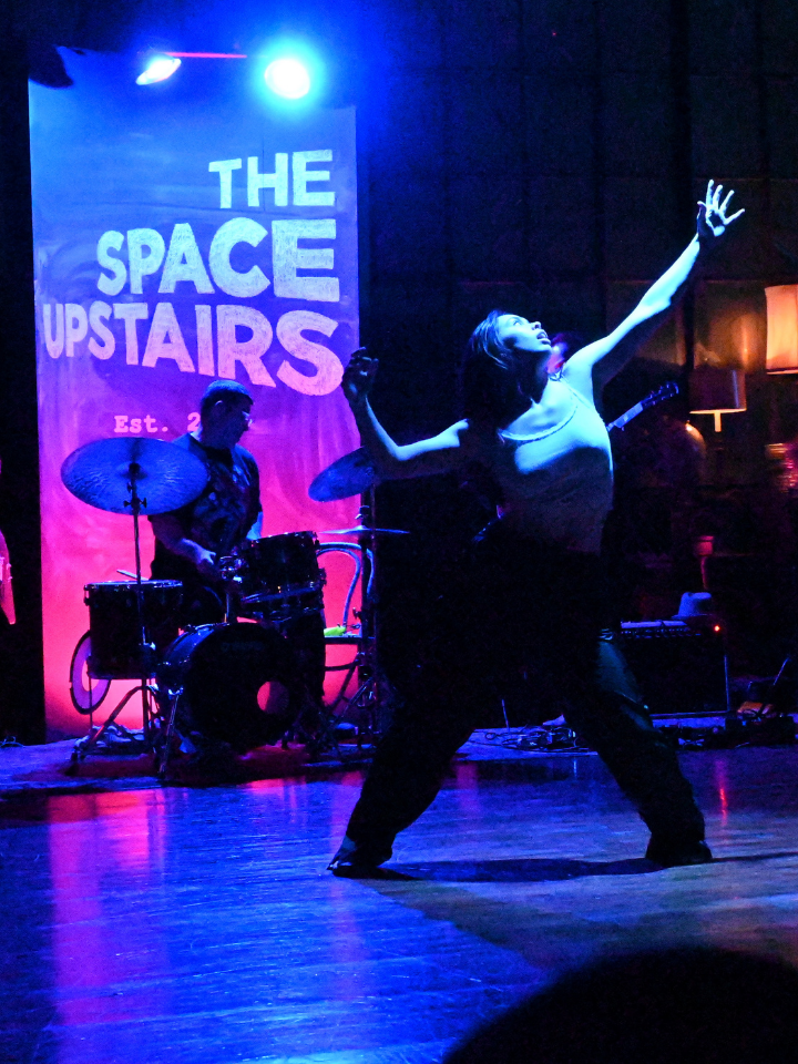 A dancer performs on stage with a band under blue and purple lighting.