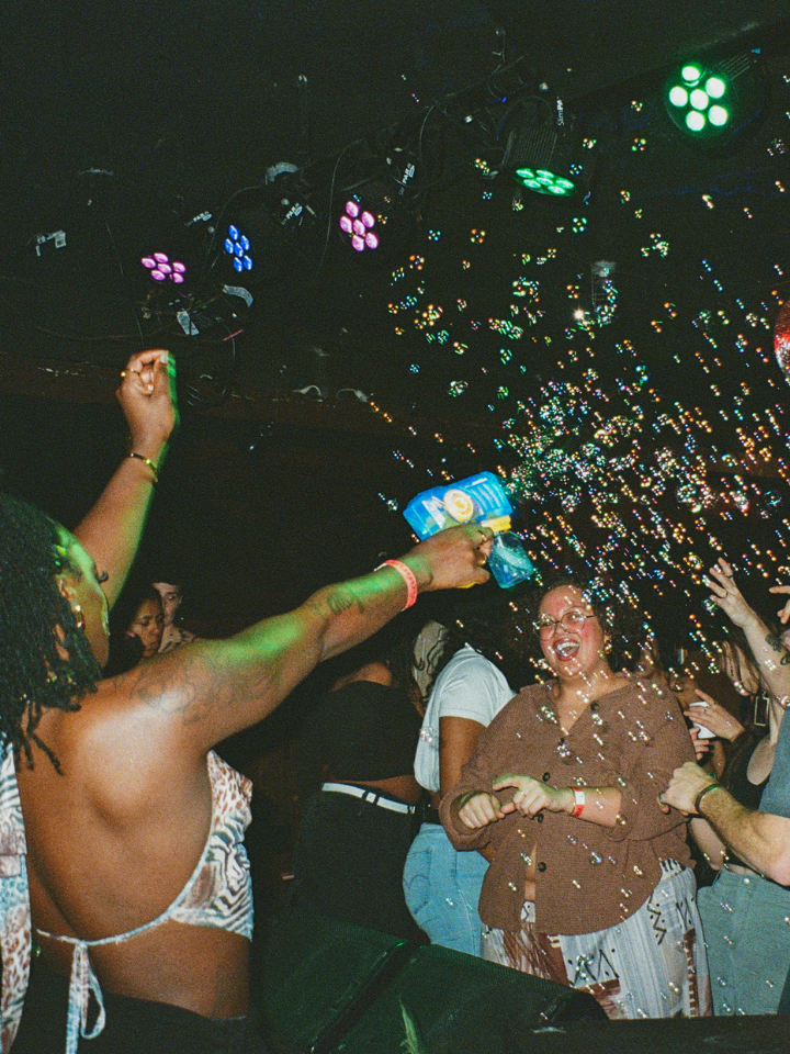 A lively crowd dances as a dancer sprays bubbles at a nightclub with colorful lights.