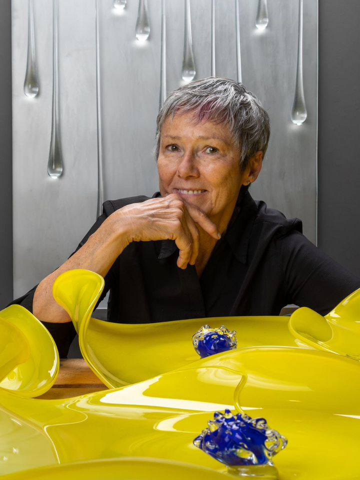 A person sits behind yellow glass art pieces, smiling with hand on chin.
