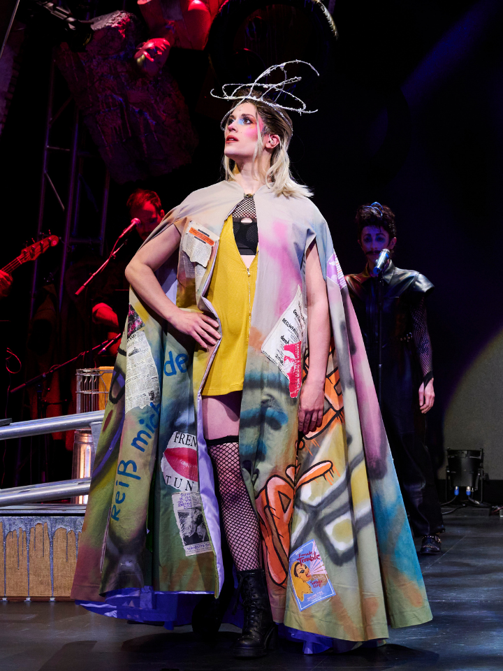Person in colorful, graffiti-style cape poses on stage with musicians and dramatic lighting.
