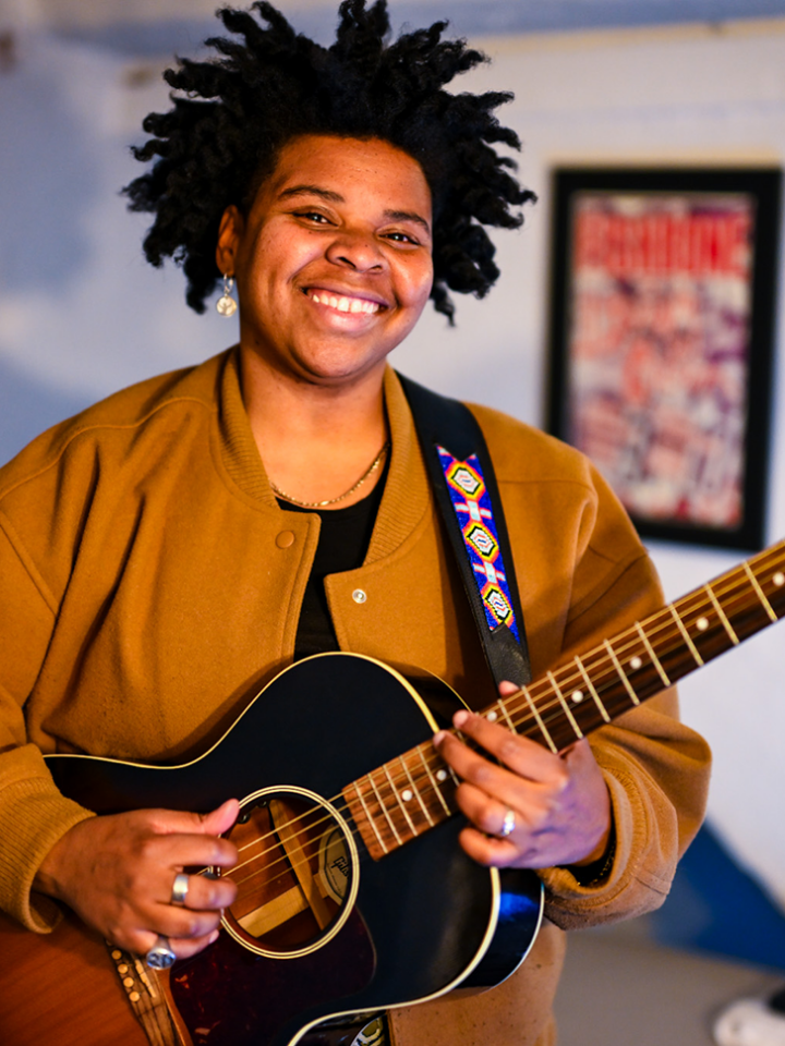 Person smiling and playing a black acoustic guitar indoors, wearing a brown jacket.