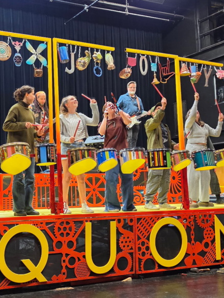 A band performs on a colorful stage with drums and hanging objects, SQUONK displayed below.