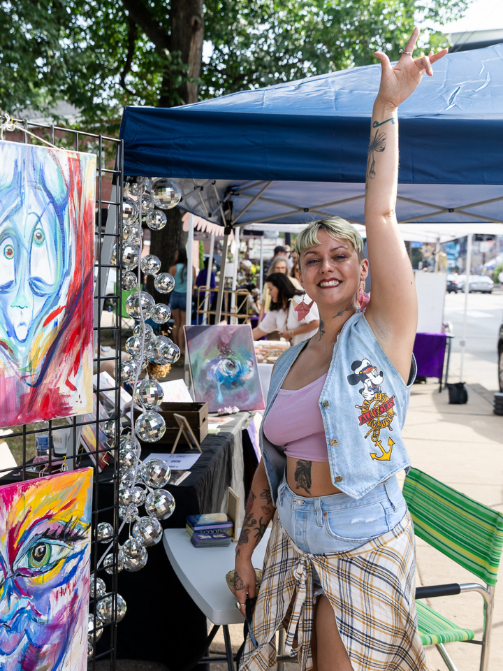 Smiling person raises arm at outdoor art booth with colorful abstract paintings and decorations.
