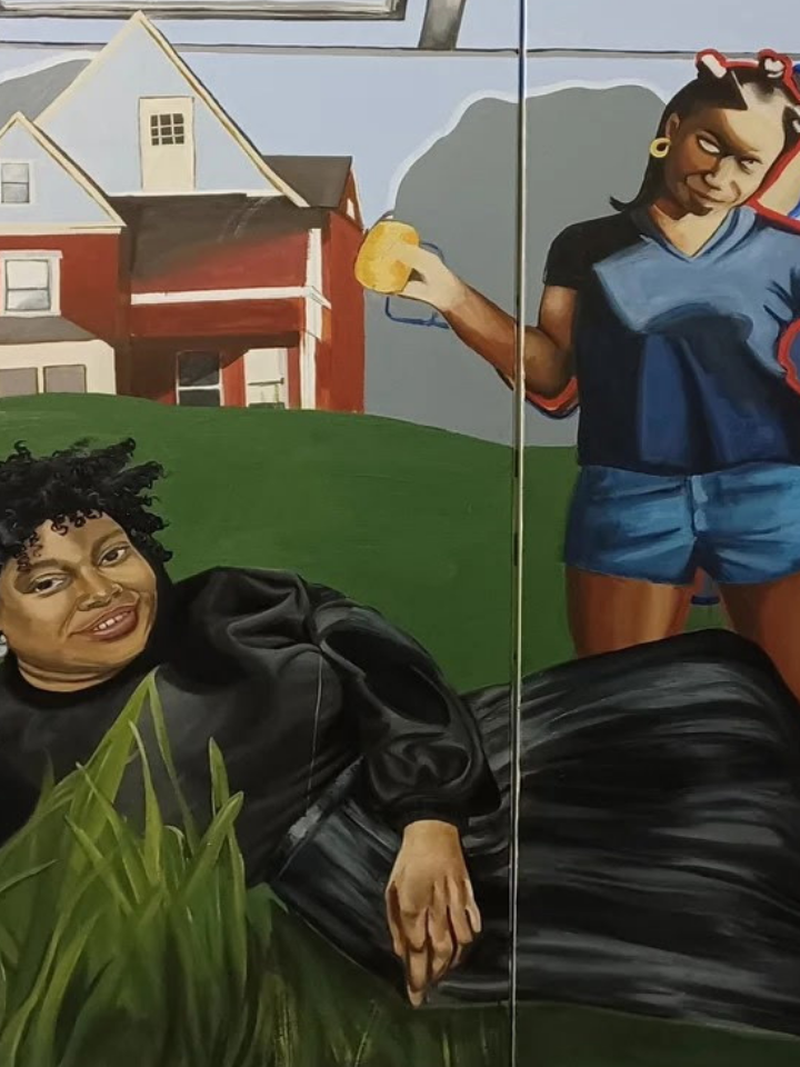 Two women pose on grass; one reclines, one stands, with a red house in the background.
