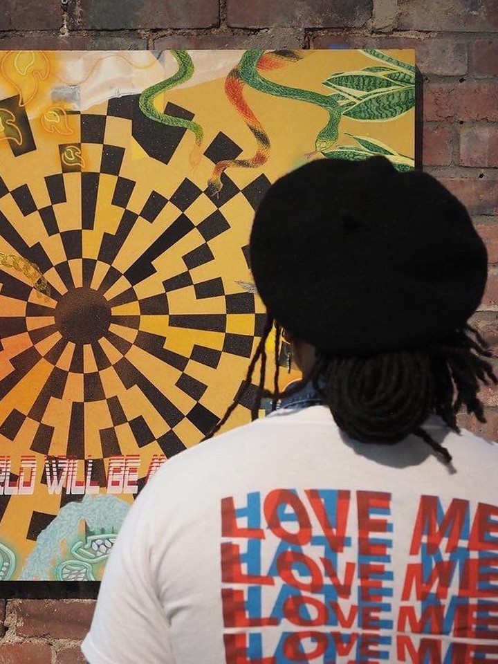 A person wearing a t-shirt that says "love me love me love me" on the back is shown from behind looking at a colorful piece of artwork hung on a brick wall