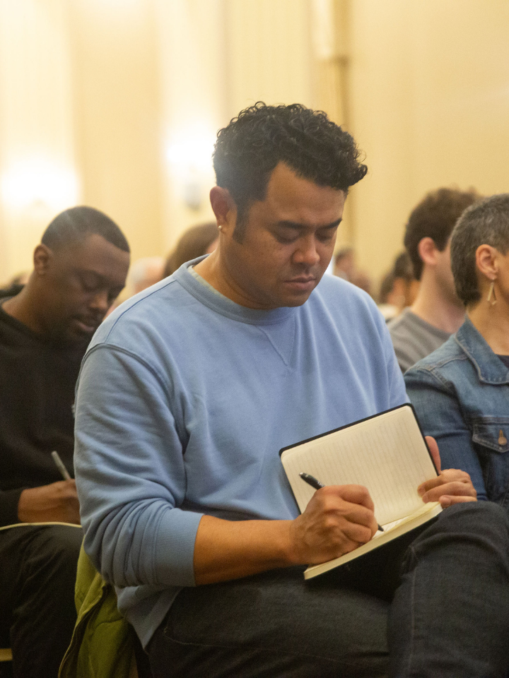 People sitting in an audience, writing in notebooks and listening attentively.