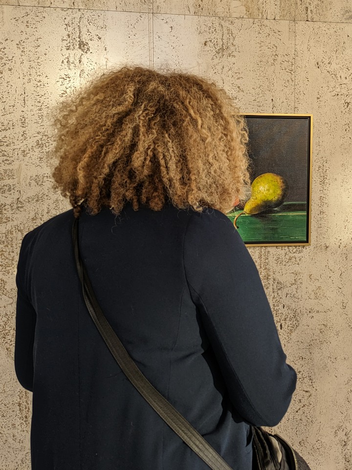 A person with shoulder-length curly light brown hair and wearing a black jacket and purse over their shoulder looks at a still life painting of a pear hanging on a wall