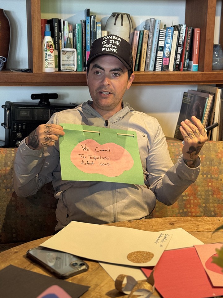 A person holds a handmade sign that reads We commit to equitable artist wages, sitting at a table with craft supplies and bookshelves in the background.