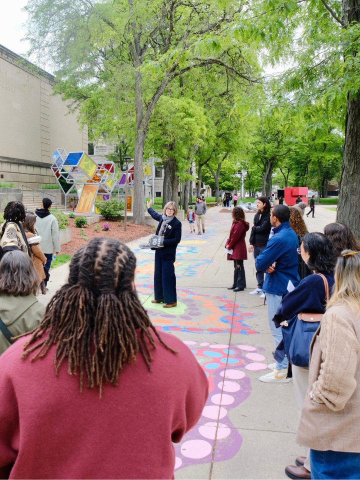 A group of people listen to a guide outdoors near a museum and colorful ground art.