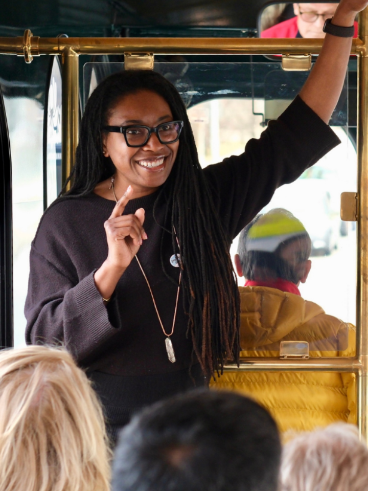 A smiling woman stands and speaks to a group of passengers at the front of a trolley