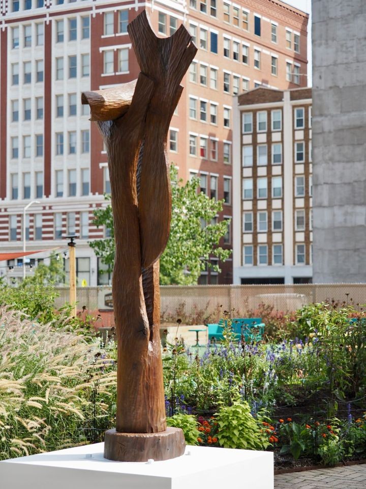 a tall sculpture that looks like it was made out of wood stands on a pedestal in front of city buildings