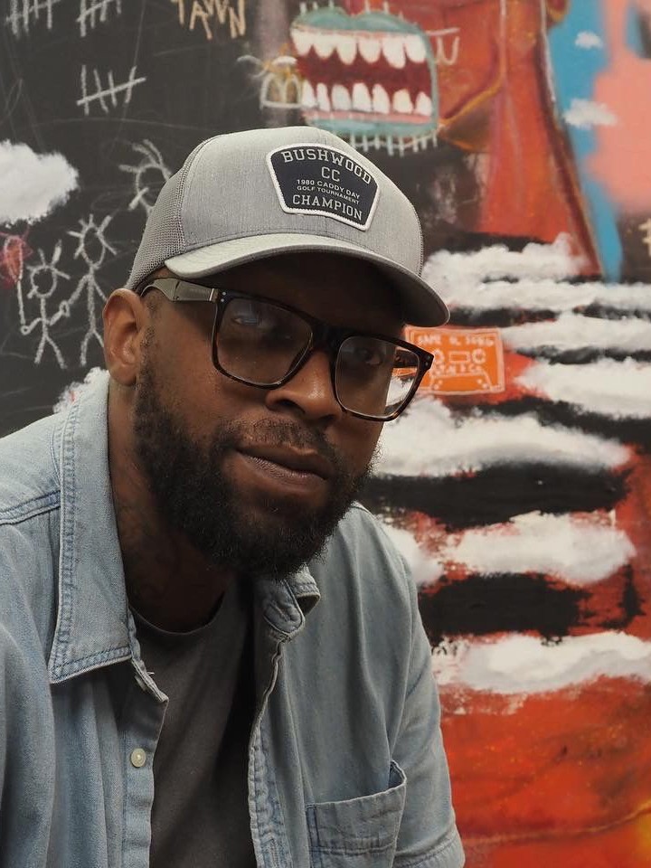 A Black man with a beard, gray baseball cap, black glasses, gray T-shirt, and denim collared shirt poses in front of a colorful painting