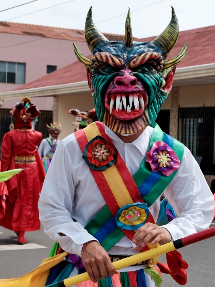 People in colorful demon masks and costumes parade down a street during a festival.