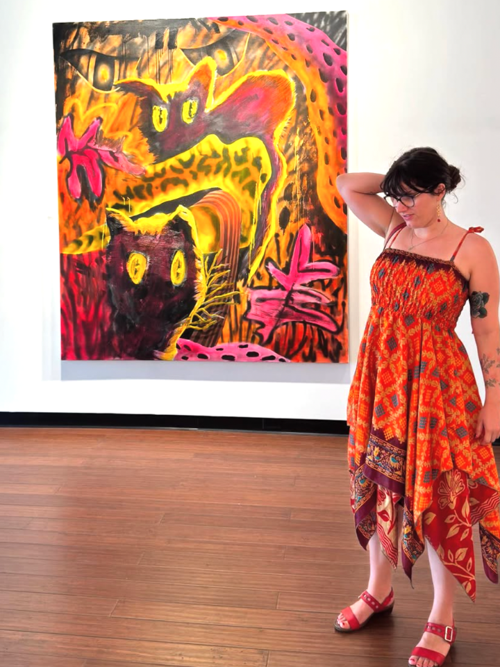Woman in a red dress stands by a colorful painting of abstract cats in a gallery.
