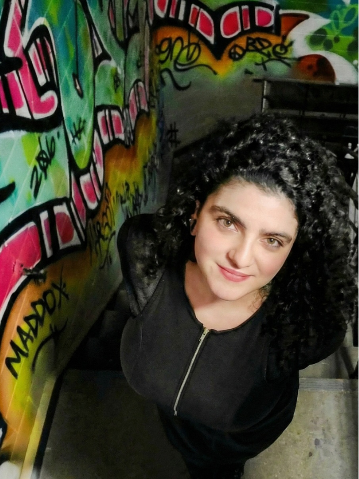 Woman with curly hair stands by graffiti-covered wall, looking up at the camera.
