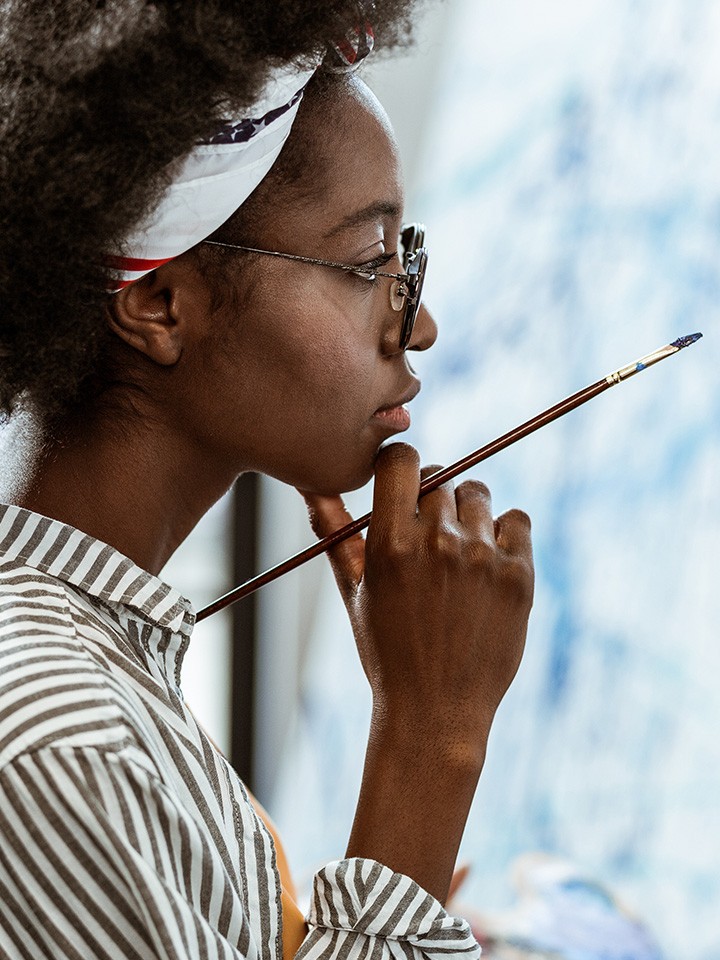 A Black woman with short hair pulled back in a headband holding a paintbrush looking off something off camera