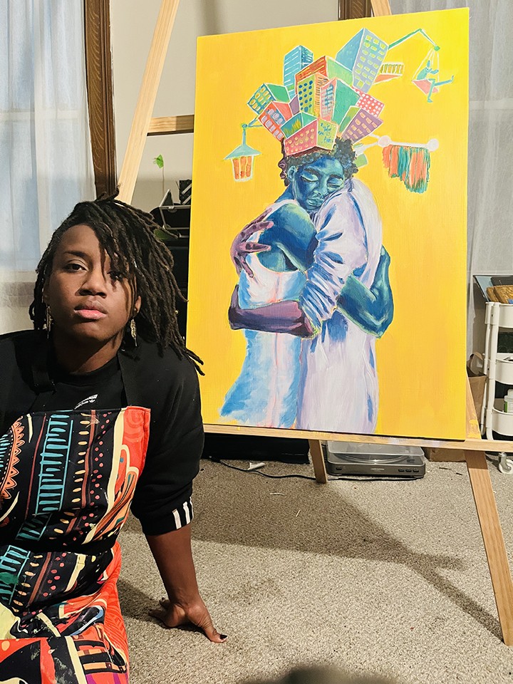 Ifeoma, a Black woman with shoulder-length dark hair wearing a black T-shirt with a colorful Africana-inspired pattern, sits in front of a colorful piece of artwork on display depicting two people hugging on a yellow background