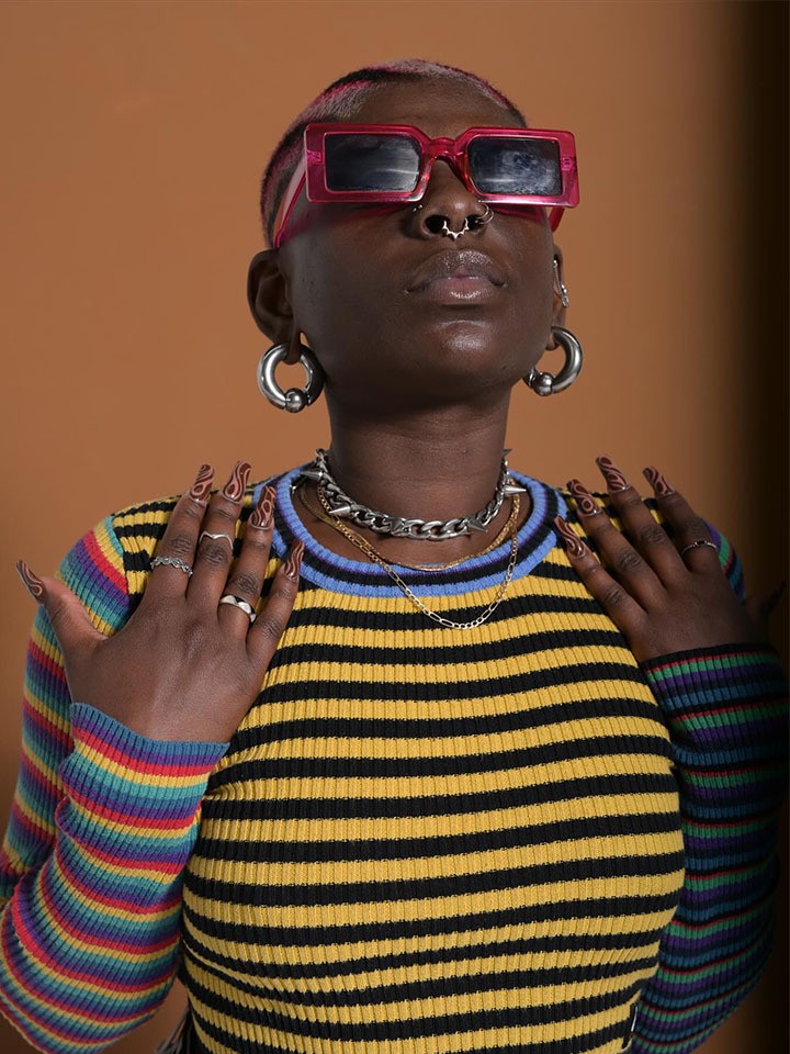 Black person with short hair, red sunglasses, large silver hoop earrings, silver necklaces, and a colorful striped long sleeved shirt