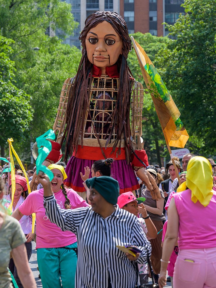 A giant 12-foot-tall puppet of a 10-year-old Syrian girl walks amid a parade of humans