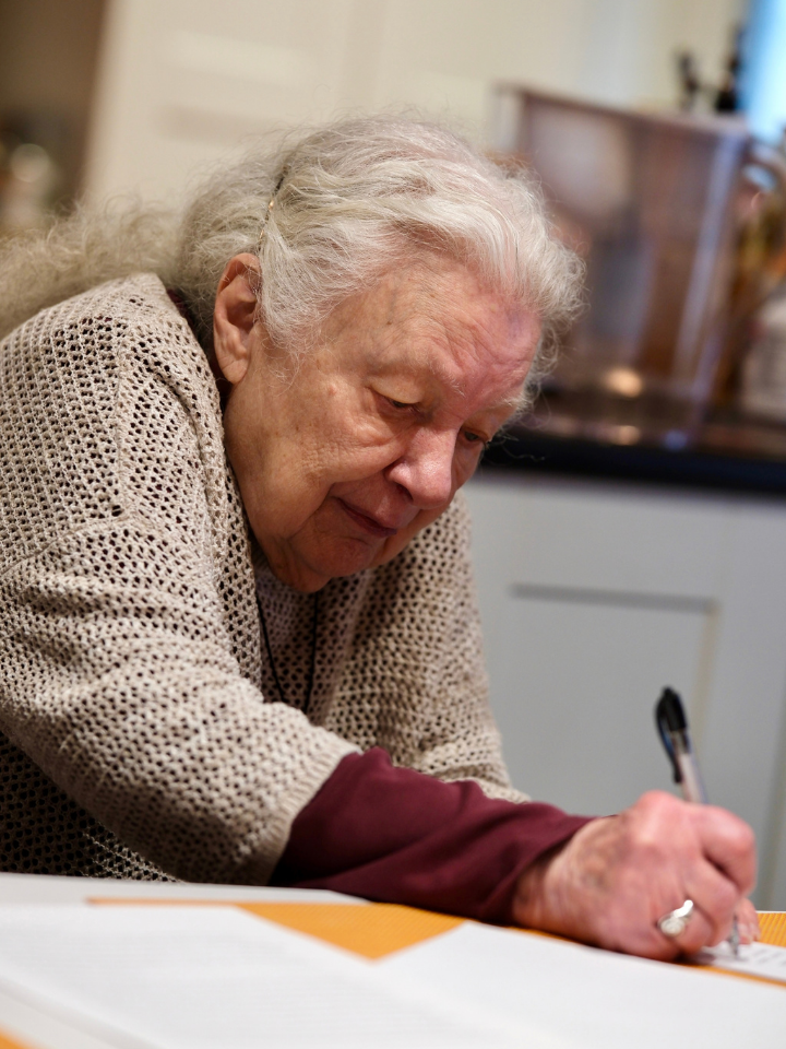An older woman with white hair writing on paper with a pen.