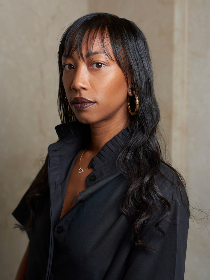 Woman with long dark hair and black shirt standing against a beige background.