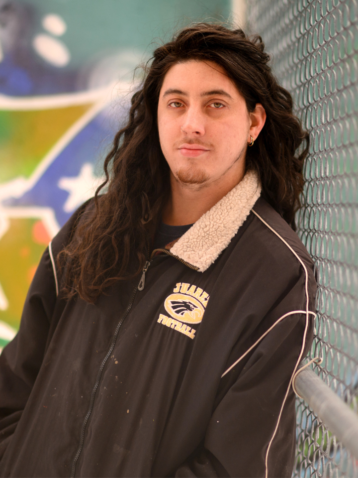 A person with long hair leans against a chain link fence outdoors.