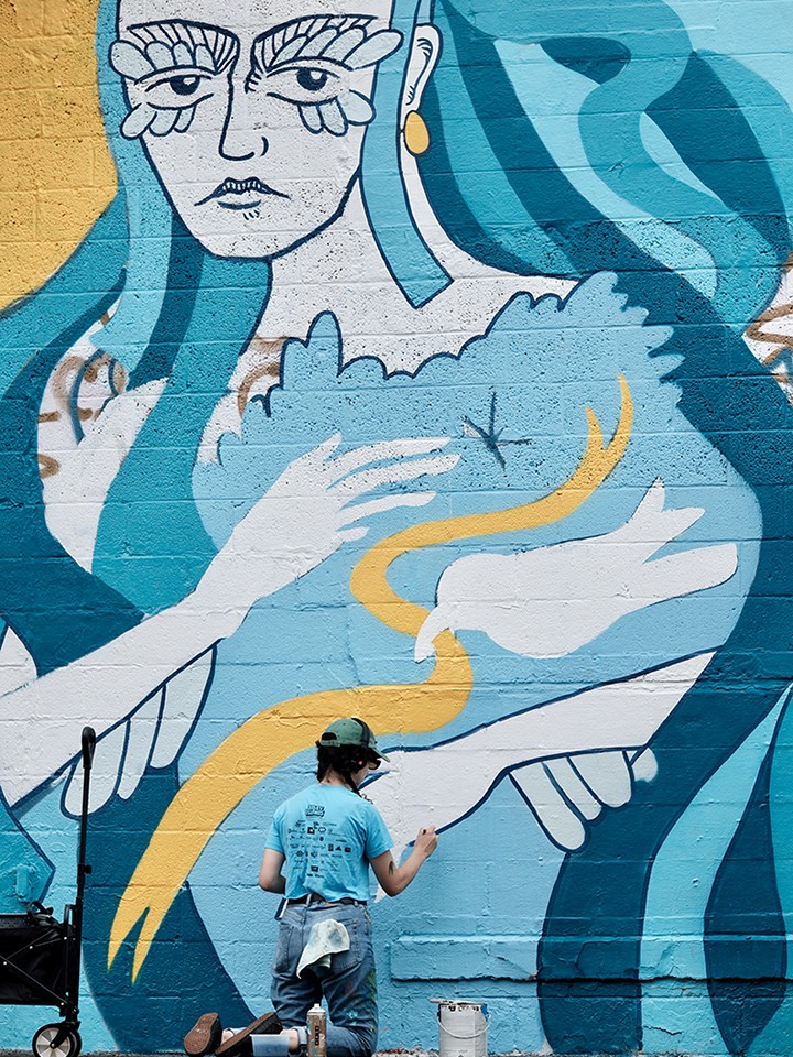 A person in a blue shirt and helmet paints a large mural of a woman with blue hair holding a white bird on a brick wall. The mural features bold lines and shades of blue, yellow, and white.