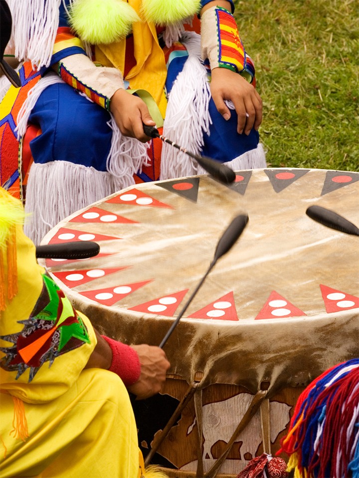 Native Americans in colorful outfits sit in a circle around a drum
