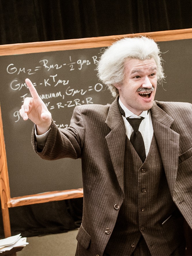 An actor portraying Albert Einstein gestures in a brown suit, with his equations drawn on a chalkboard behind him.