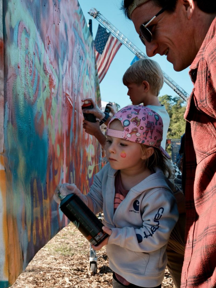 A young child in a pink cap spray paints a colorful mural while a man watches.