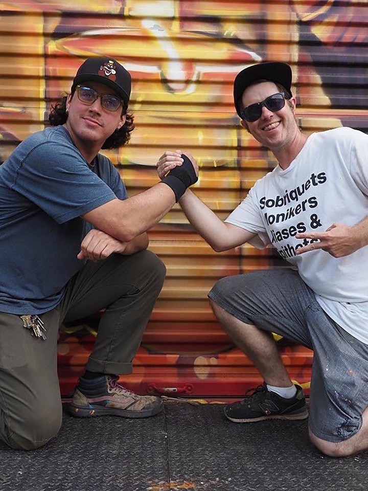 Two men grip hands and smile for the camera while posing in front of a graffiti mural painted on a metal door