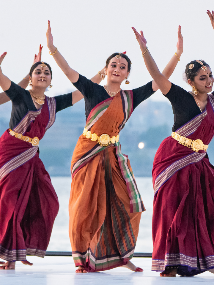 Three women perform a traditional Indian dance in colorful sarees and gold jewelry.