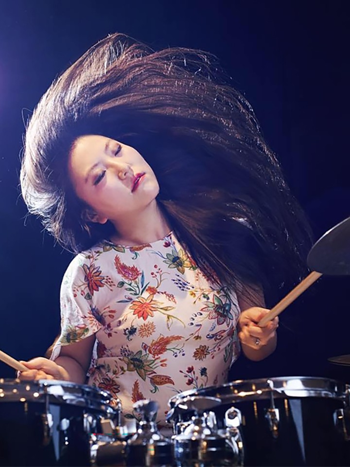 An Asian woman with long dark straight hair and wearing a white shirt with a floral pattern closes her eyes and tilts her head as she plays drums