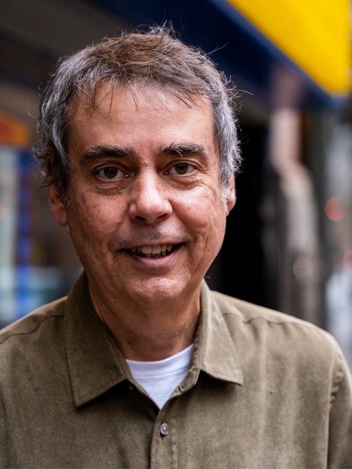 Portrait of a smiling man with short gray hair, a white T-shirt, and a light brown collared button-down shirt. He's standing outside in front of a blurred background that shows a glimpse of a theater marquee.