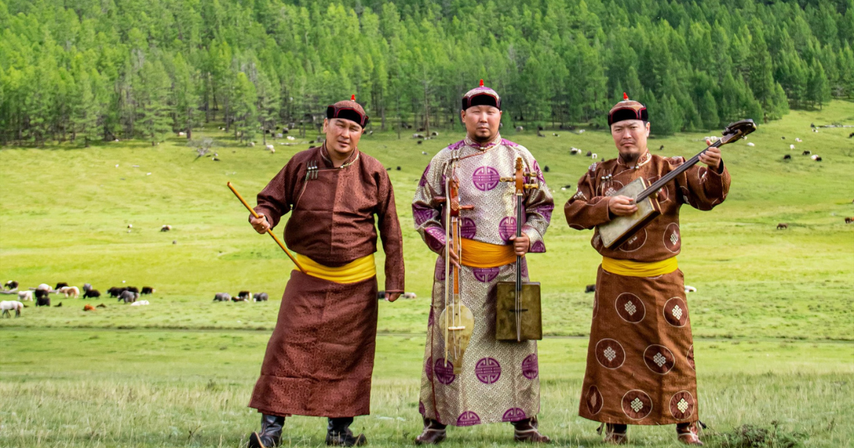 Three men in traditional Mongolian clothing stand on a grassy field with trees and grazing animals in the background. One holds a stringed instrument, and all wear hats and bright yellow sashes.