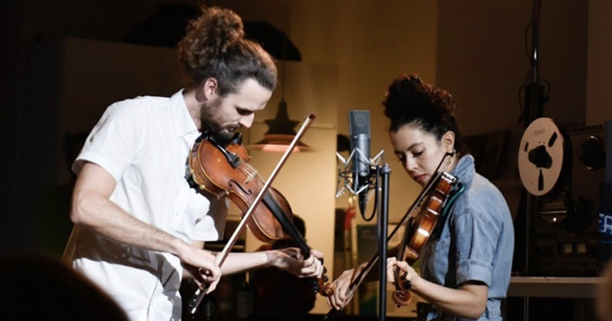 Two people playing violins in front of a microphone in a dimly lit room.